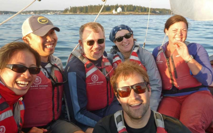 Six people wearing life jackets sit in a sailboat and smile at the camera. The water is blue and calm.